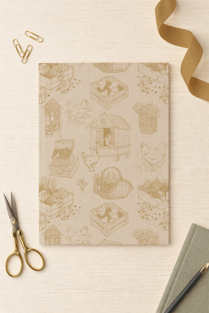 A mockup of a notebook with the pattern Hens & Garden delight, designed by Martie Erasmus for Creative Faith Studio. This pattern forms part of the Rooted by Grace collection featuring golden illustrations of hens and farm life on a cream background. Patterns available for licensing.