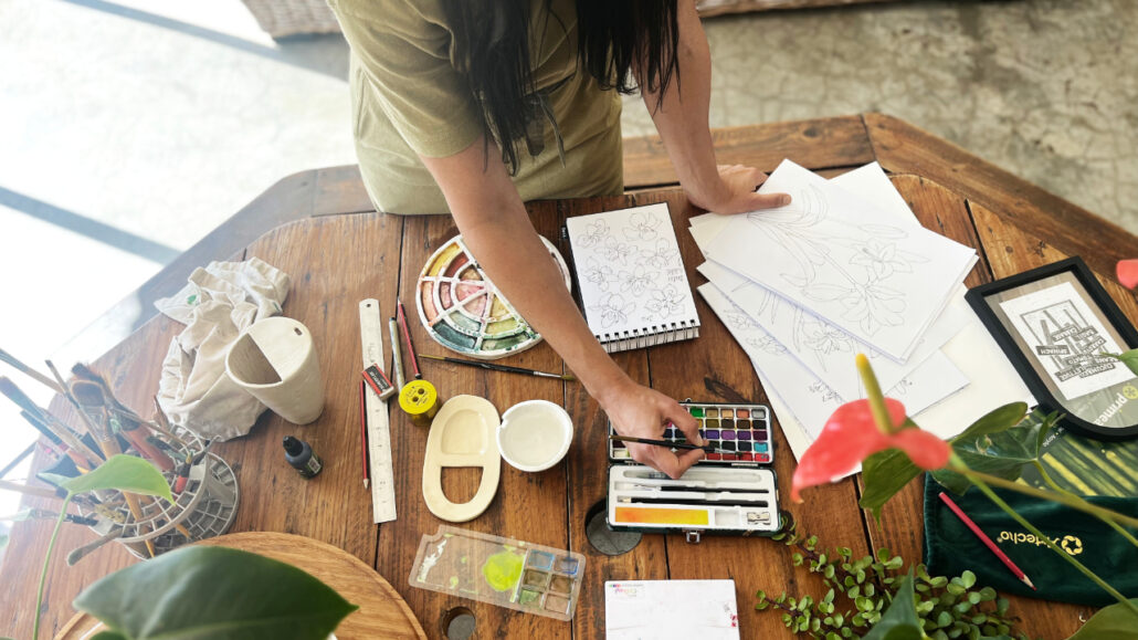 Surface Pattern Designer, Martie Erasmus standing at a wooden table with paintbrush and watercolour palette. The table is full of inspirational plants and art supplies as she creates artwork for her Seamless repeat patterns for art licensing.