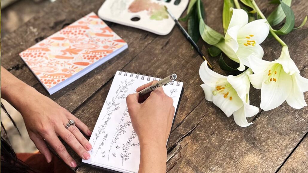 Artist Martie Erasmus drawing with pen in her skectbook laid out on a wooden table with a notebook, watercolour palette and white lily flowers while creating motifs for her Surface Pattern Designs for Art Licensing.