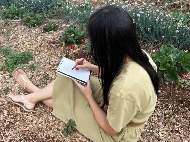 Artist Martie Erasmus in a green dress sitting in her strawberry garden with a sketchbook and drawing while creating artwork for her Seamless Repeat Patterns.