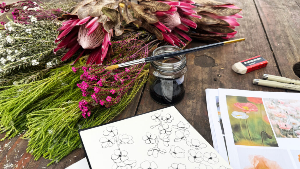 Cosy creative workspace with sketchbooks, ink, protea and other fynbosbotanical references, and unfinished artwork bathed in natural sunlight.
