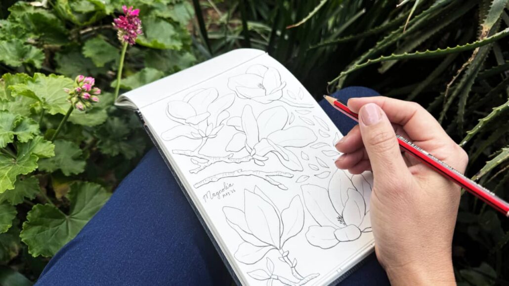 Open sketchbook of artist Martie Erasmus resting on her lap in a lush garden surrounded by flowers and greenery, representing creativity rooted in faith and abundance.